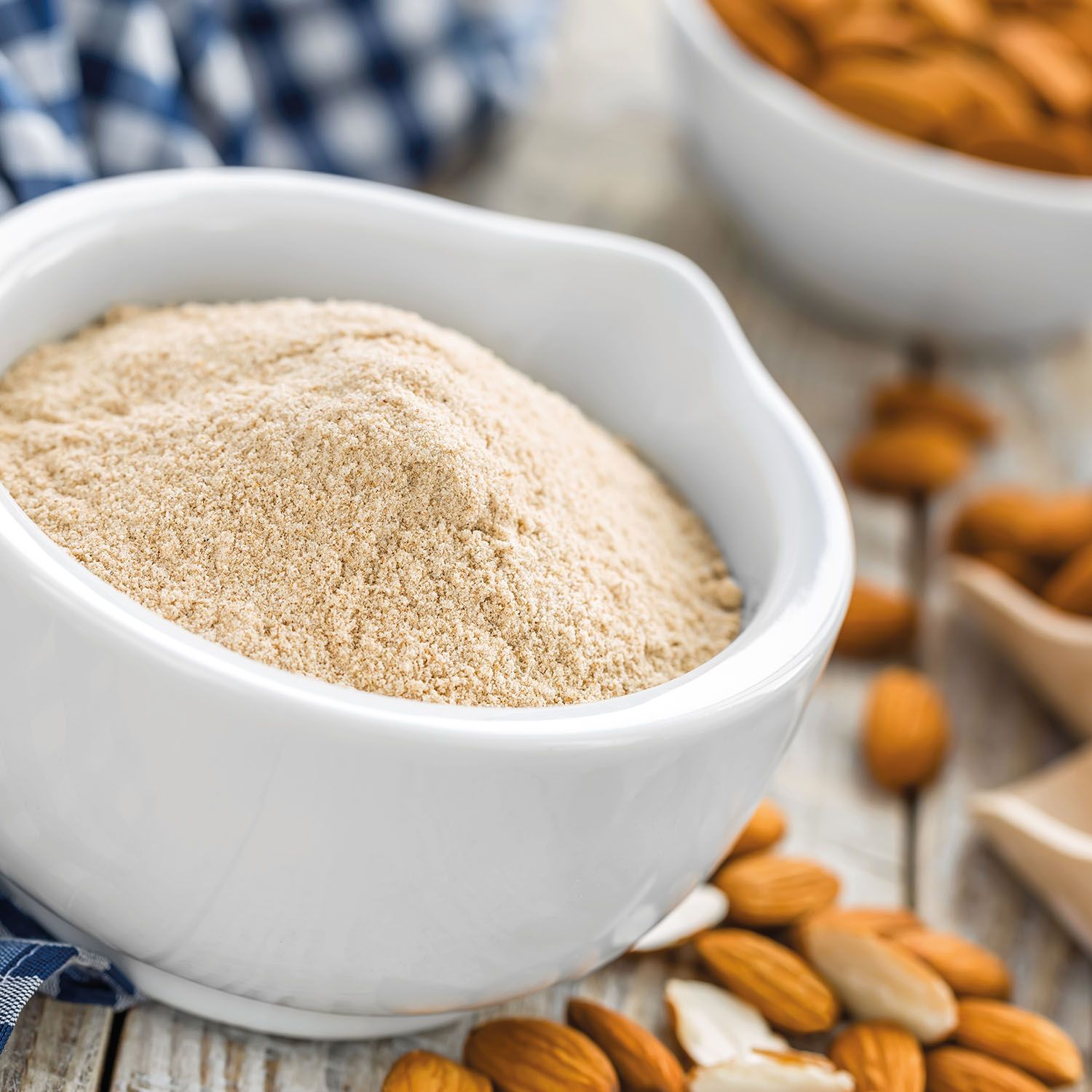 Weißes Mandelmehl in einer weißen Schüssel. Daneben Mandeln. Blaue Serviette im Hintergrund.