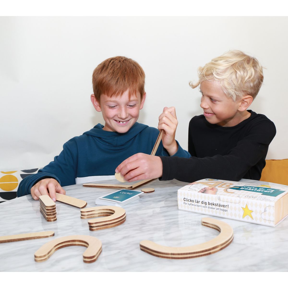 Zwei Kinder am Tisch. Holzbuchstaben, Karten und Box. Kind hält Holzteil. Kind hält Holzteil. Buchstaben U und S.