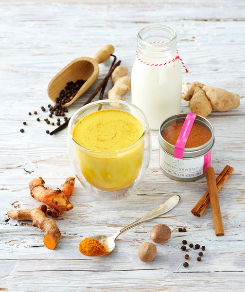 Glas mit goldener Milch, Zutaten: Kurkuma, Ingwer, Zimt, Pfeffer, Muskatnuss. Dose mit Gewürz. Holzuntergrund.