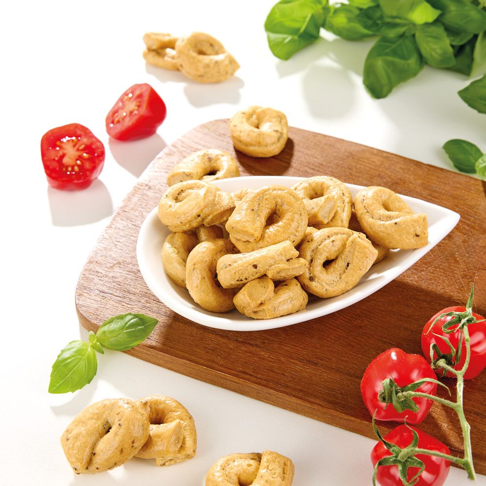 Taralli-Gebäck auf weißem Teller, Tomaten und Basilikumblätter auf Holzbrett.