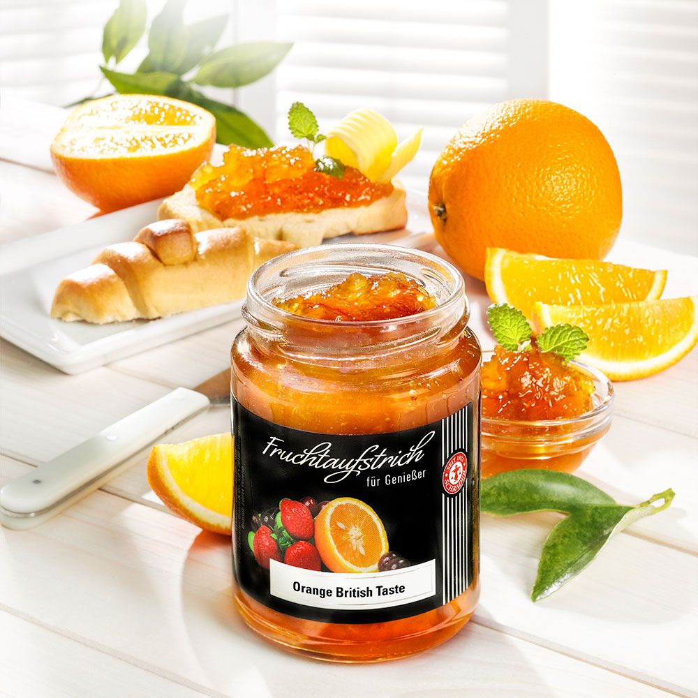 Glas Orangenmarmelade, daneben Brot mit Marmelade, Orangenstücke und Blätter. Etikett "Orange British Taste".