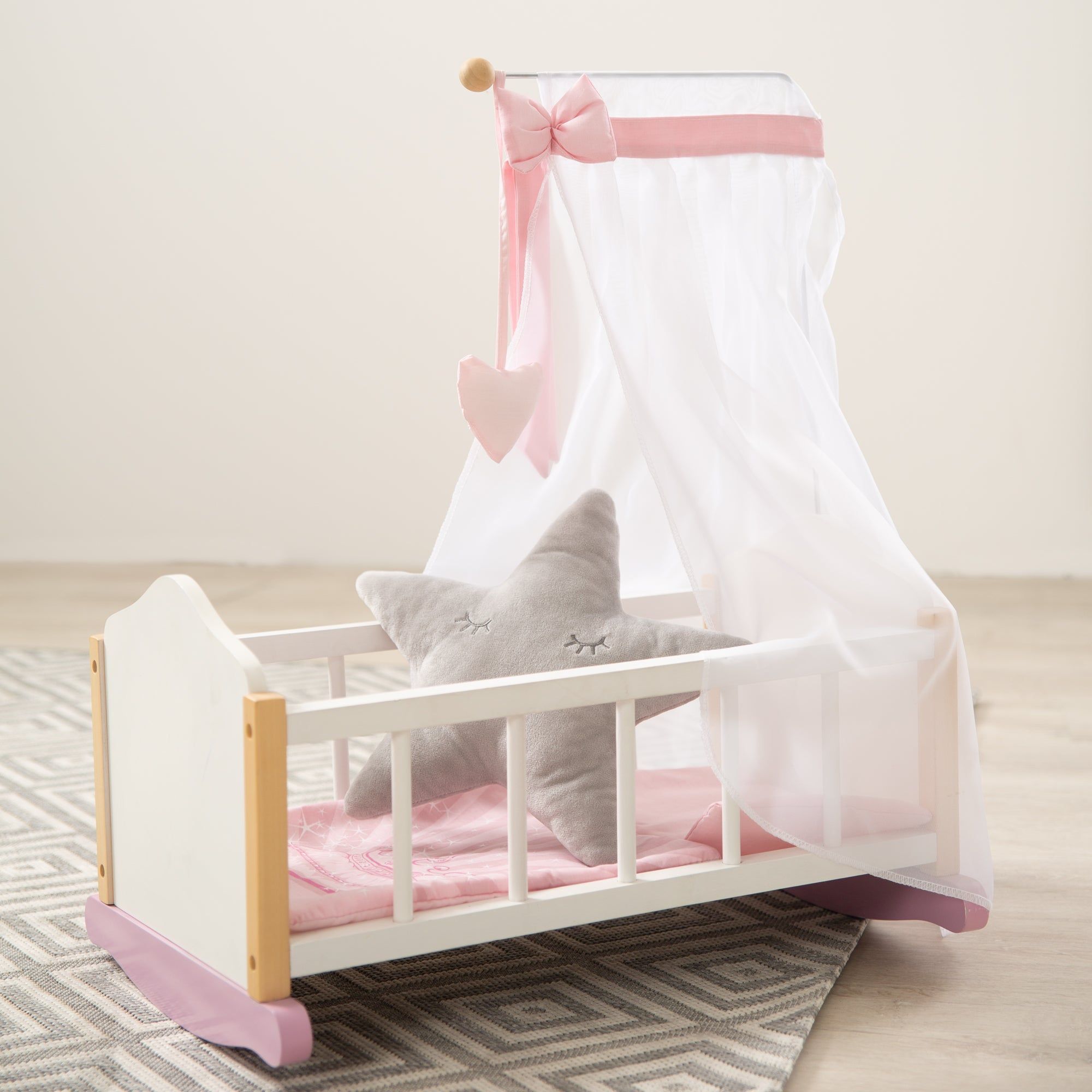 Puppenwiege mit Baldachin, Sternkissen und rosa Bettwäsche. Auf einem Teppich.