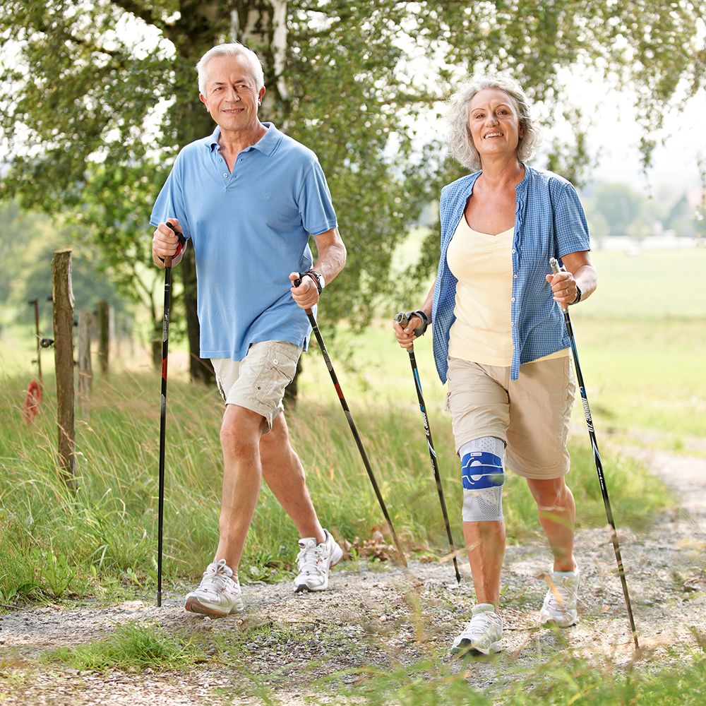 Älteres Ehepaar beim Spaziergang. Frau mit Kniebandage GENUTRAIN A3 NATUR LINKS 1. Beide mit Wanderstöcken.