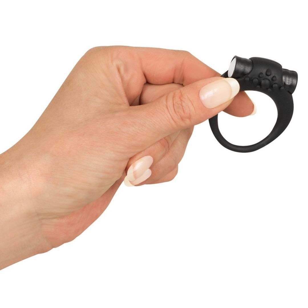 Schwarzer Penisring, gehalten von einer Hand. Ring mit Noppen und zwei zylindrischen Elementen.