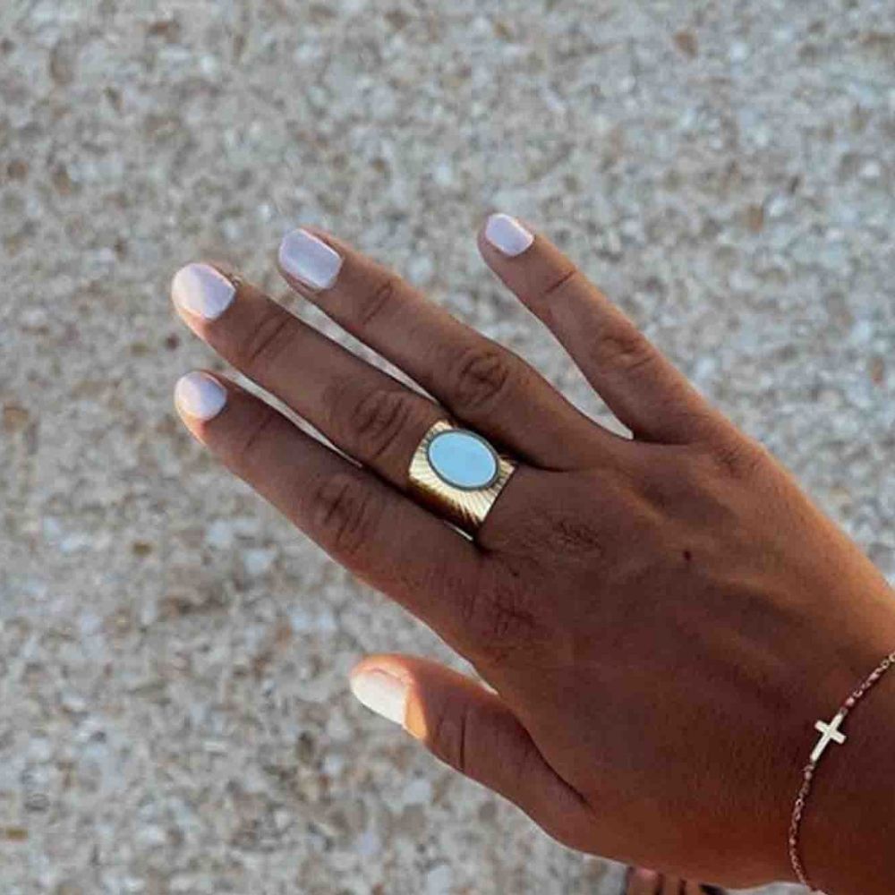 Hand mit hellgrau lackierten Fingernägeln, trägt einen goldenen Ring mit blauem Stein und ein Armband.