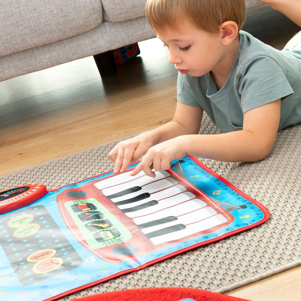 Ein Kind liegt auf einem Teppich und spielt auf einer Musikmatte mit Klaviertasten. Ein roter Knopf ist sichtbar. Ein Sofa ist im Hintergrund.
