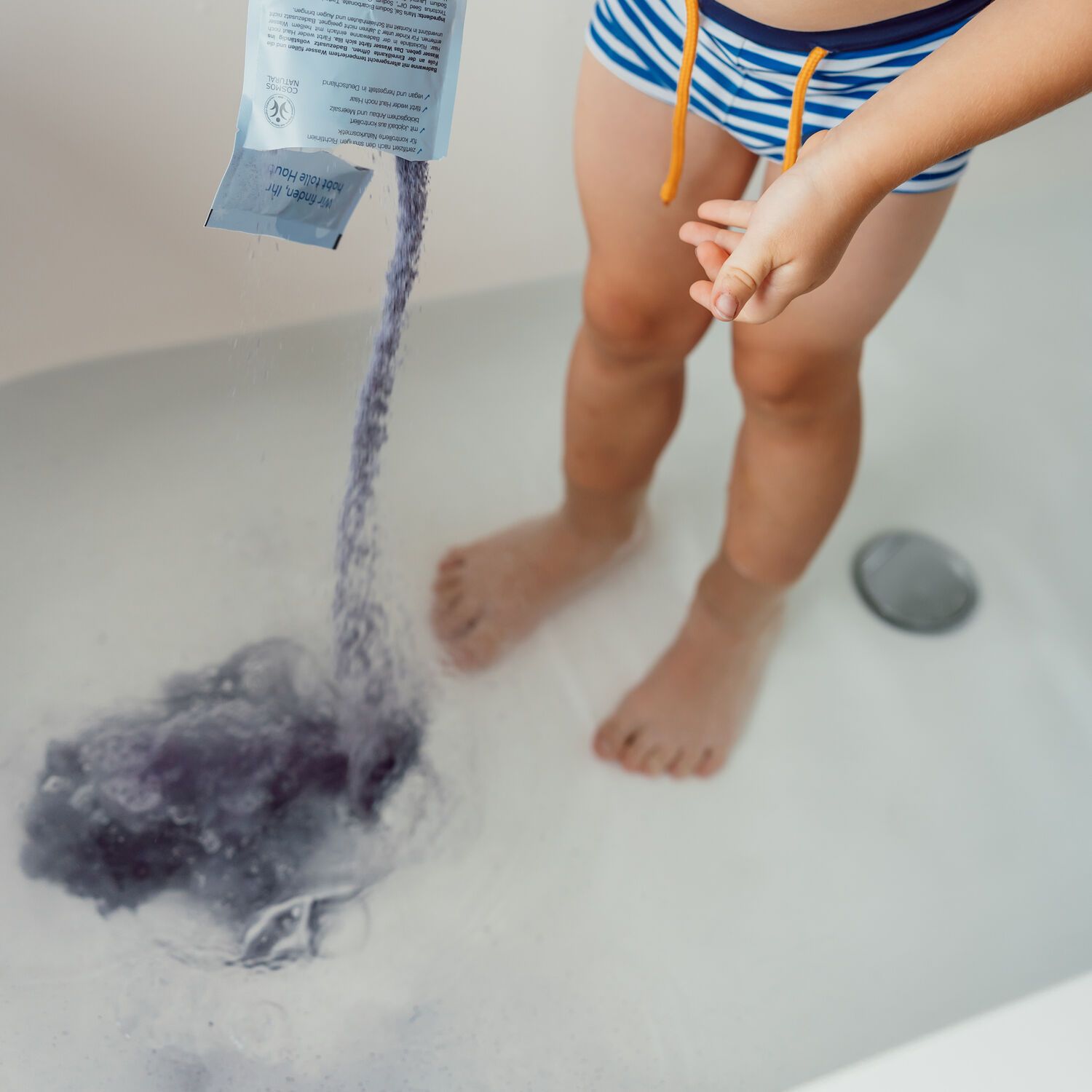 Kind in Badehose steht in Badewanne. Aus Beutel wird violettes Badepulver ins Wasser gegossen. Wasser färbt sich violett.