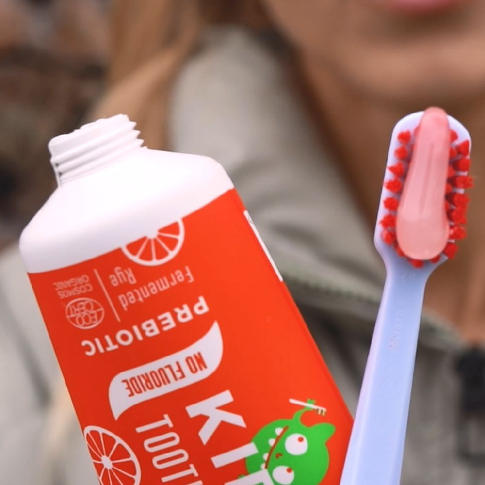 Orange Tube mit weißem Verschluss und Zahnbürste. Aufschrift: KIDS TOOTHPASTE, PREBIOTIC, NO FLUORIDE. Mit Illustrationen.