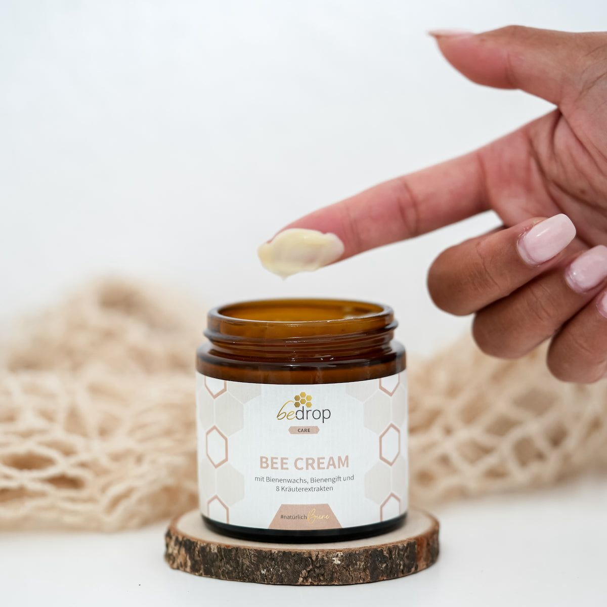 Braune Glasdose "bedrop Bee Cream" auf Holzscheibe. Finger mit Creme. Hintergrund unscharf.