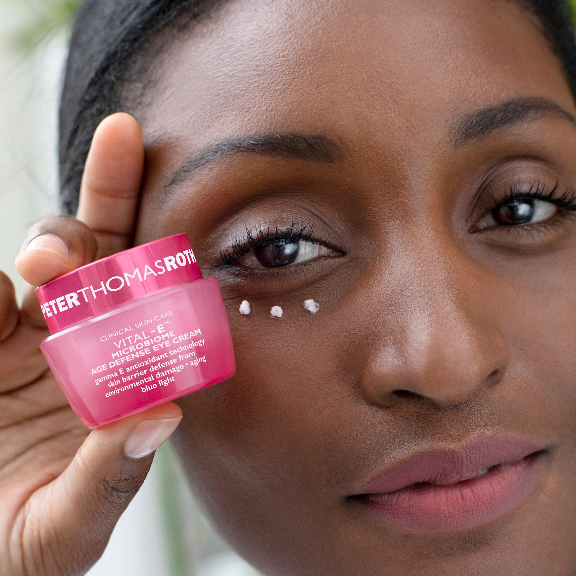 Frau hält rosa Tiegel. Creme-Punkte unter dem Auge. Text: VITAL-E MICROBIOME AGE DEFENSE EYE CREAM. Aufschrift PETERTHOMASROTH.