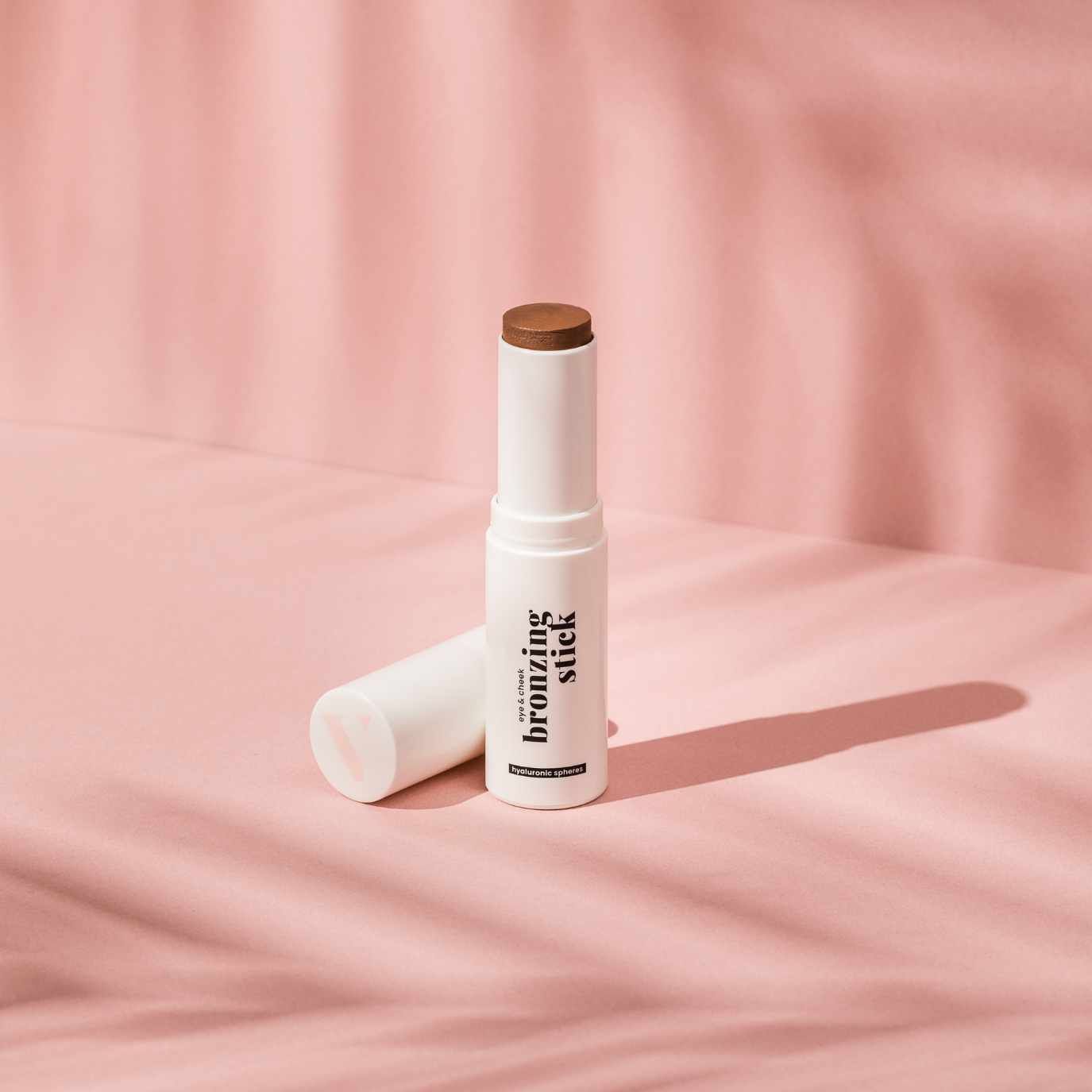 Weißer Bronzing Stick mit brauner Spitze und schwarzer Schrift auf rosa Hintergrund. Deckel liegt daneben.