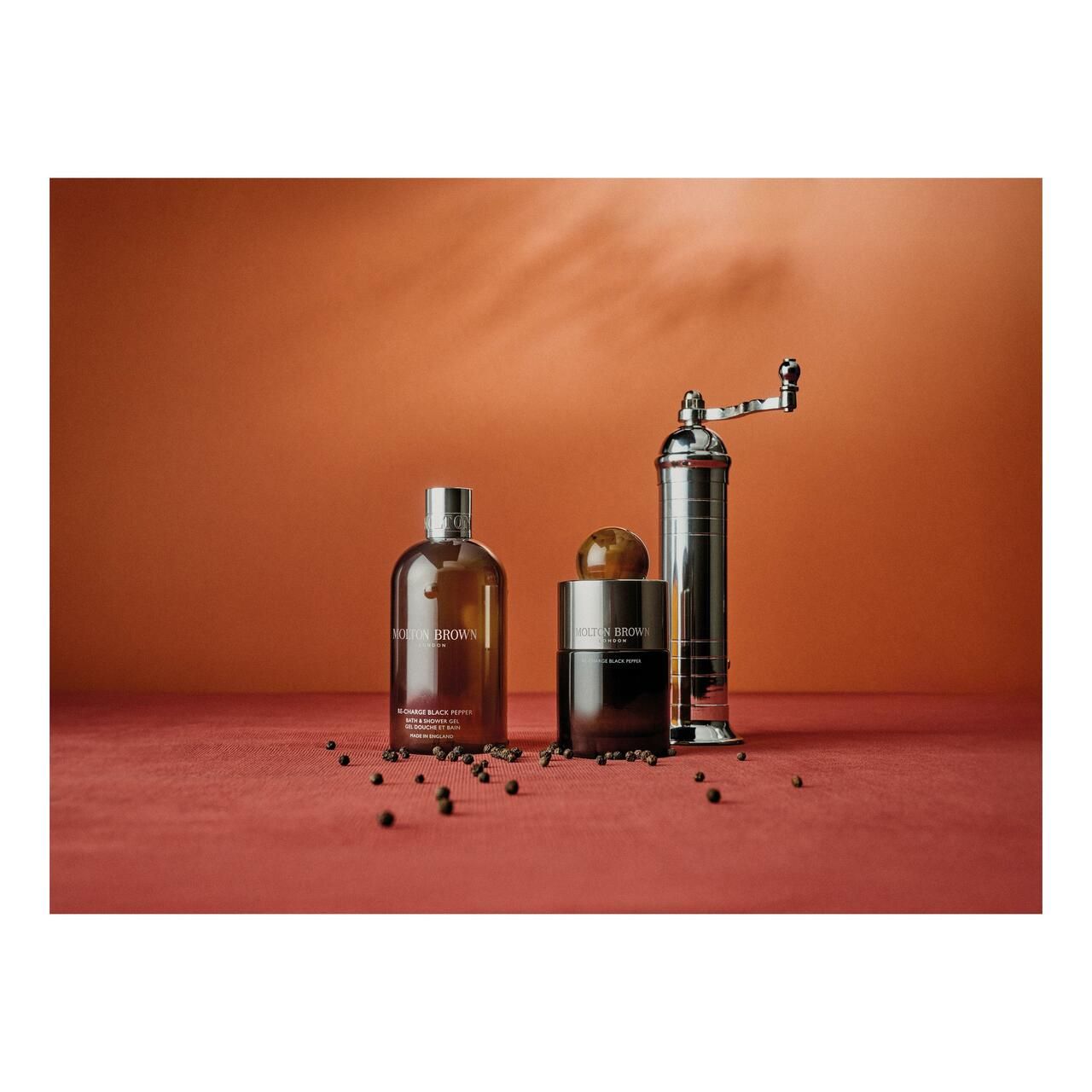 Drei Produkte und eine Pfeffermühle auf rotem Untergrund. Aufschrift: Molton Brown. Schwarze Pfefferkörner verstreut.