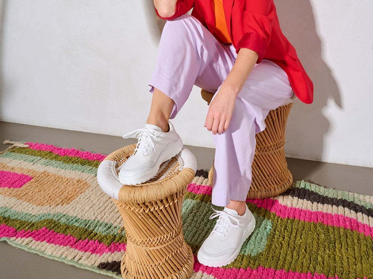 Person sitzt auf einem Hocker. Trägt weiße Schnürschuhe. Bunte Kleidung. Auf einem bunten Teppich.