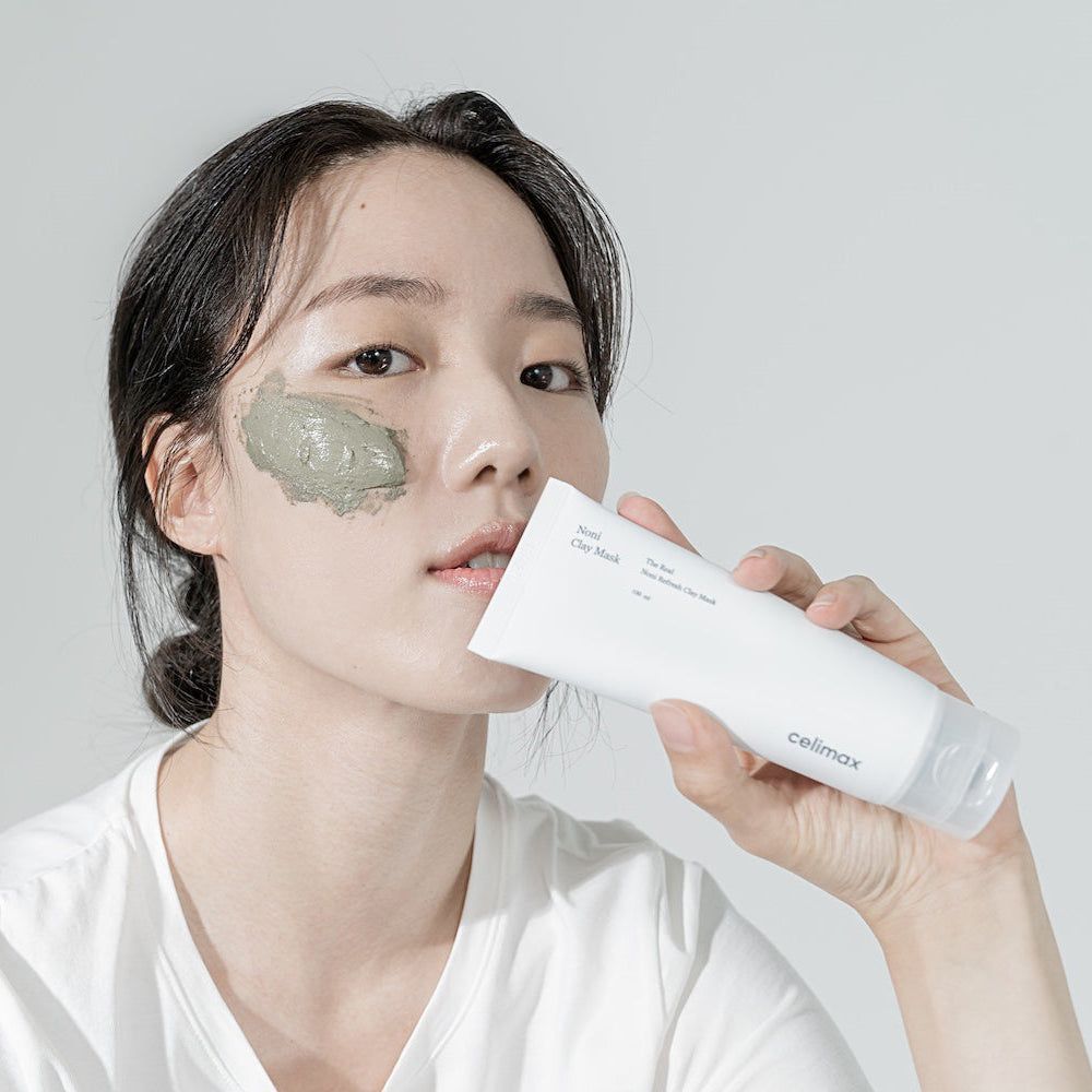 Frau trägt eine grüne Tonmaske auf, hält eine Tube CELIMAX "The Real Noni Refresh Clay Mask".