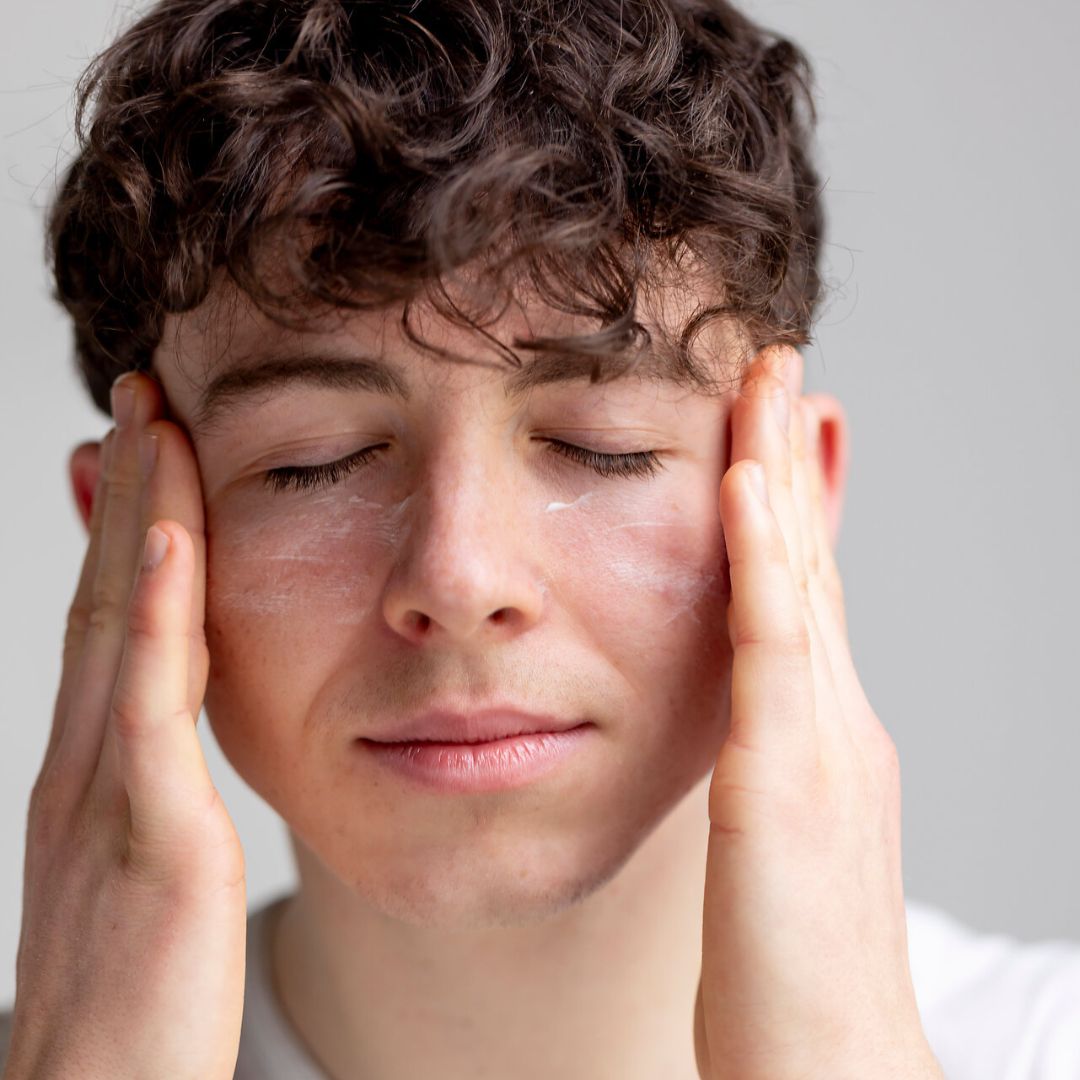 Mann mit Creme im Gesicht. Hände an den Wangen. Augen geschlossen. Creme auf der Haut. Hintergrund grau.