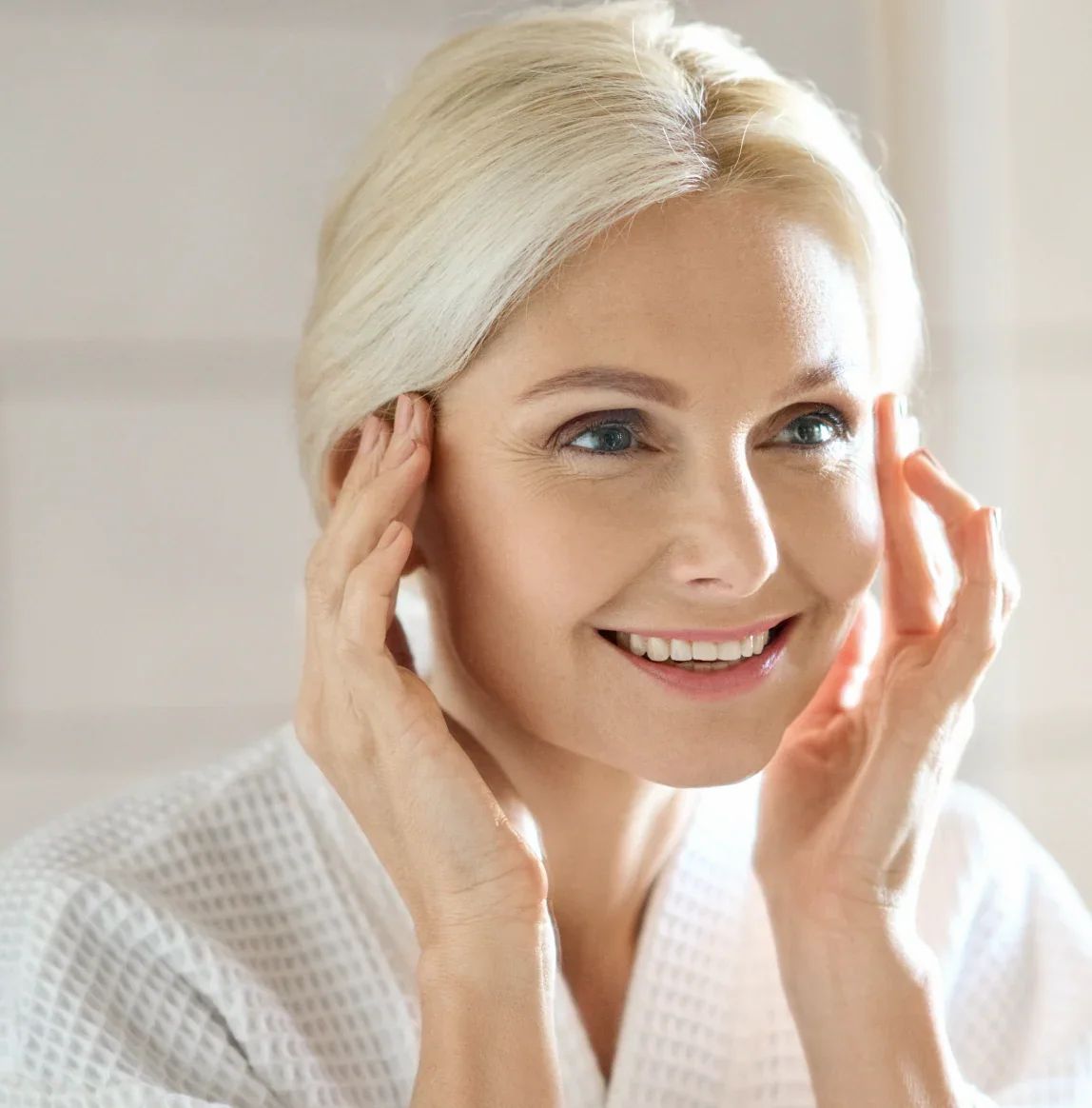 Porträt einer Frau mit blonden Haaren, die sich die Augenpartie berührt. Sie lächelt.