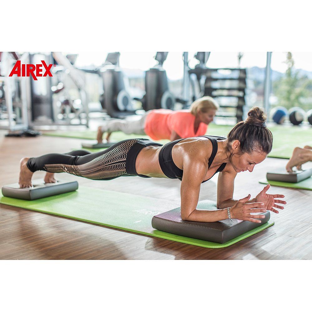 Frau trainiert im Fitnessstudio auf einer Airex Matte und einem grauen Kissen. Airex Logo.