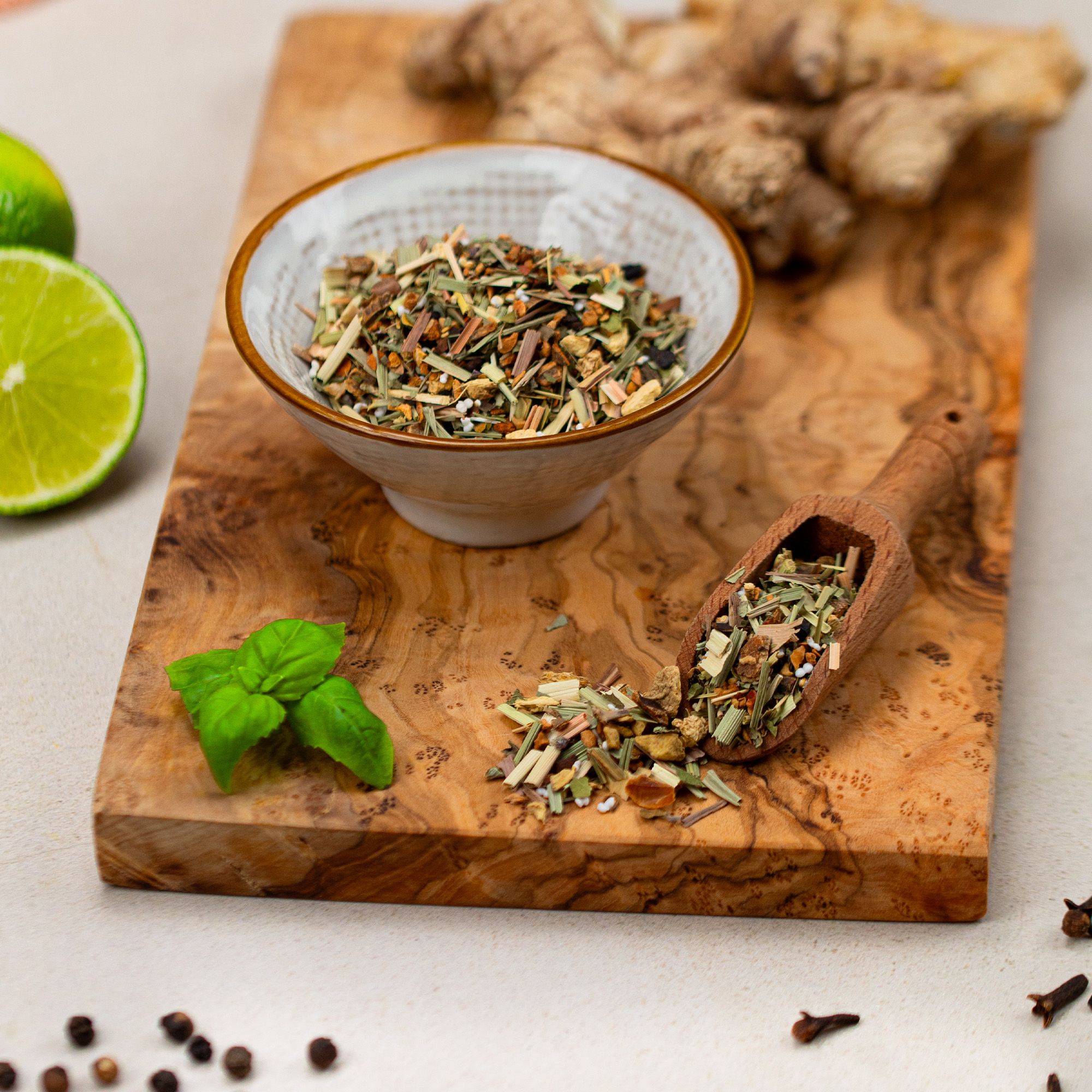 Schale mit Kräutertee-Mischung, Holzlöffel, Limette, Holzbrett. Ingwer und Gewürze daneben.