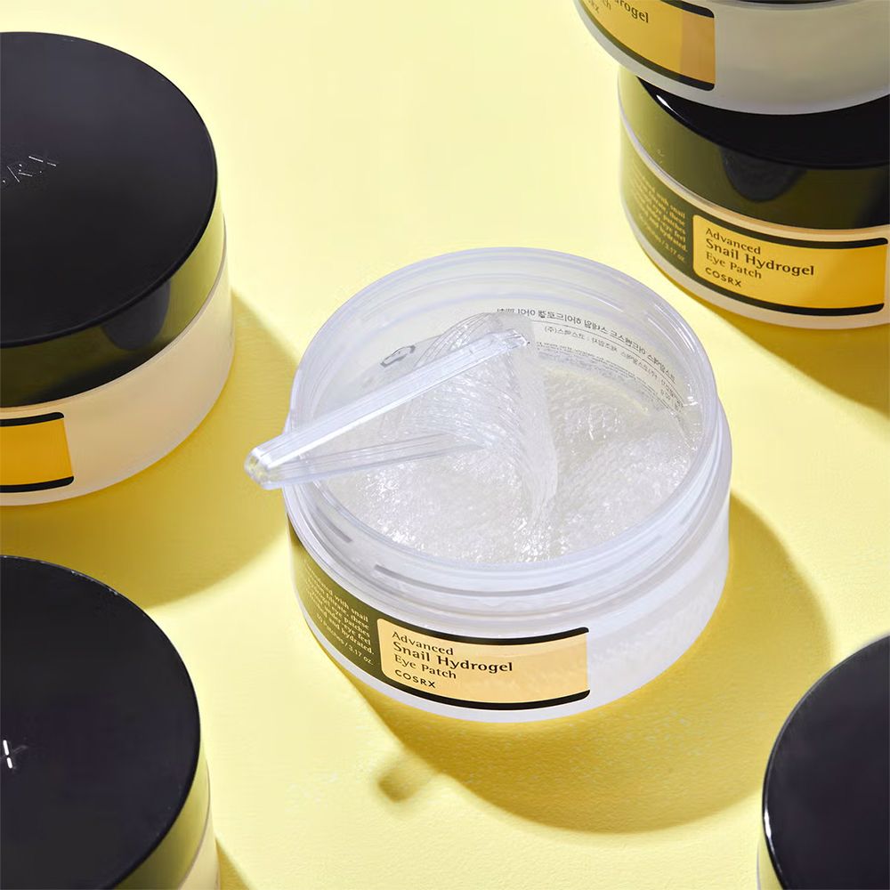 Ein offener Behälter mit Hydrogel-Augenpads und einem Spatel. Mehrere geschlossene Behälter sind im Hintergrund sichtbar.