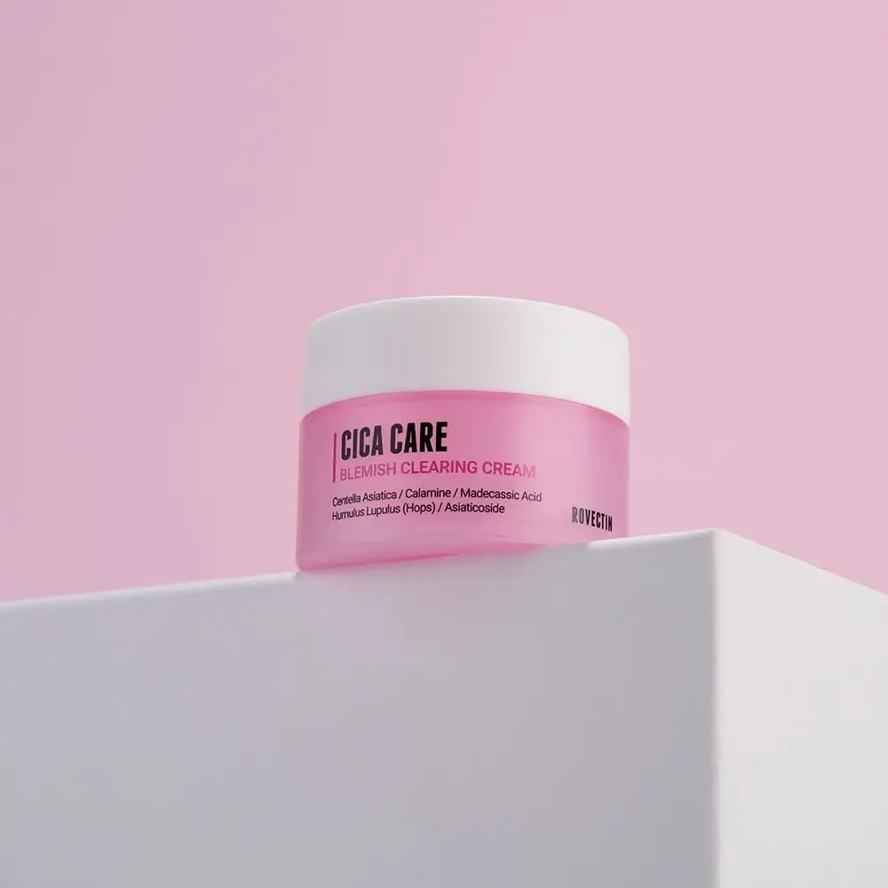Rosa Creme-Dose mit weißem Deckel. Aufschrift: Cica Care Blemish Clearing Cream. Marke: Rovectin. Auf weißer Oberfläche vor rosa Hintergrund.