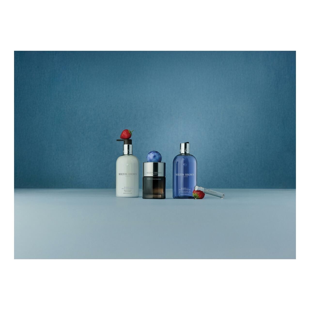 Drei Flaschen auf blauem Hintergrund. Eine Flasche mit Aufschrift Molton Brown, Bluebell & Wild Strawberry. Dekoration mit Früchten.