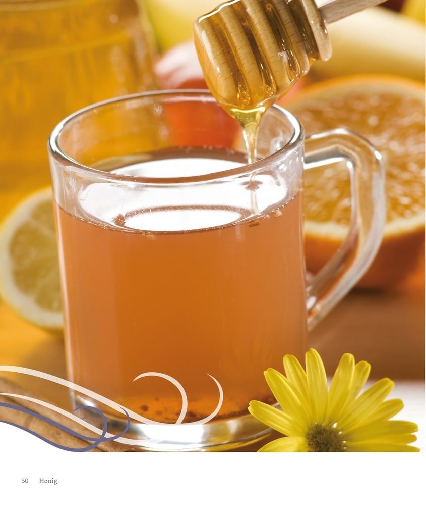 Glas mit Honig und Flüssigkeit. Honiglöffel taucht ein. Gelbe Blume und Zitrusfrüchte im Hintergrund.