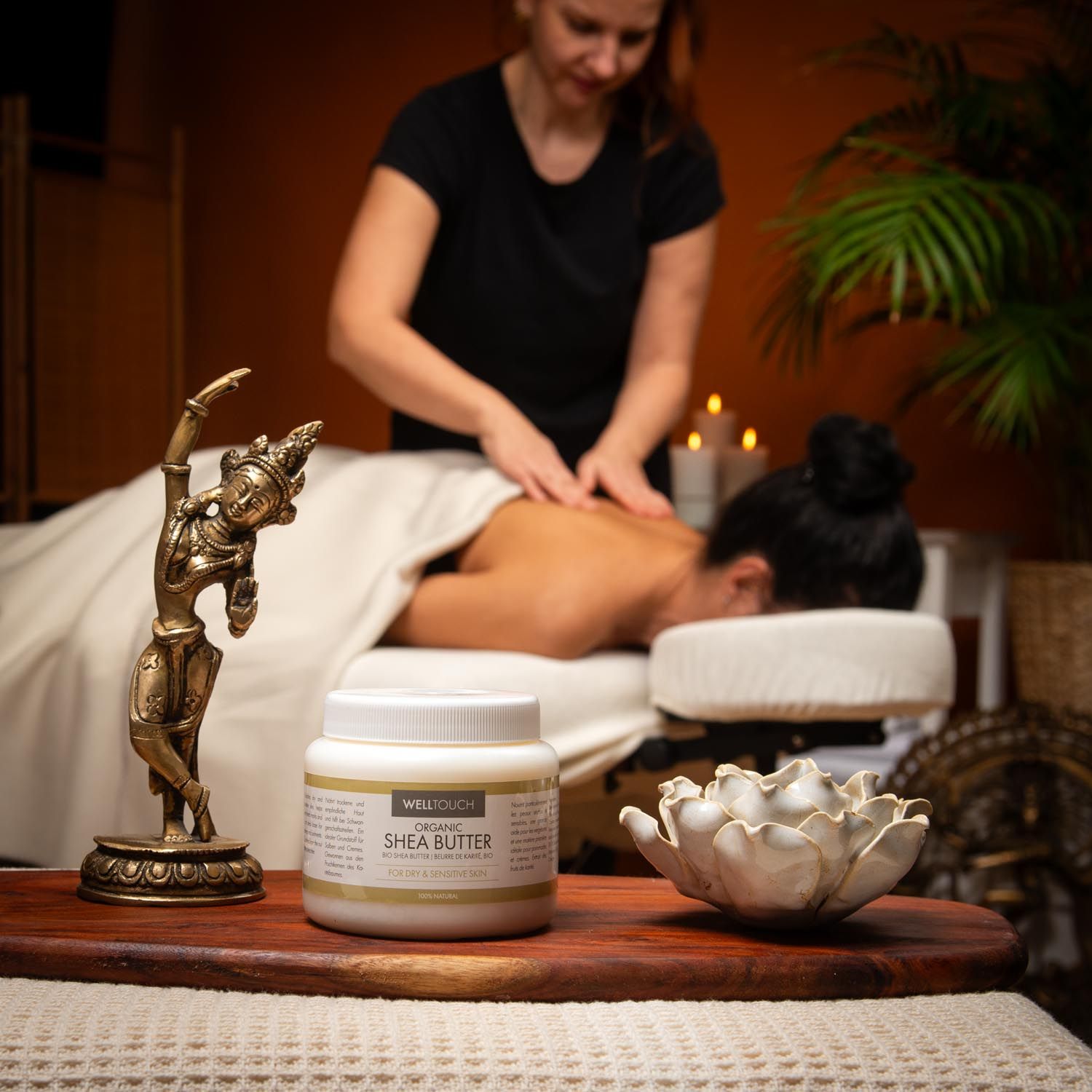 Weißes Glas Welltouch Sheabutter auf Holztablett. Frau massiert Person auf Massageliege. Goldene Figur und Lotusblüte daneben.