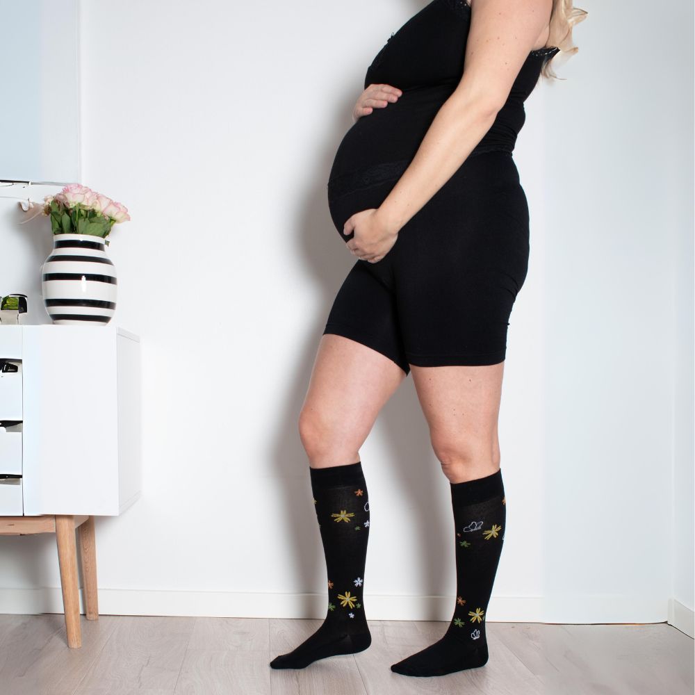 Schwangere Frau mit schwarzen Shorts und Stützstrümpfen mit Blumenmuster. Marke SupCare sichtbar.