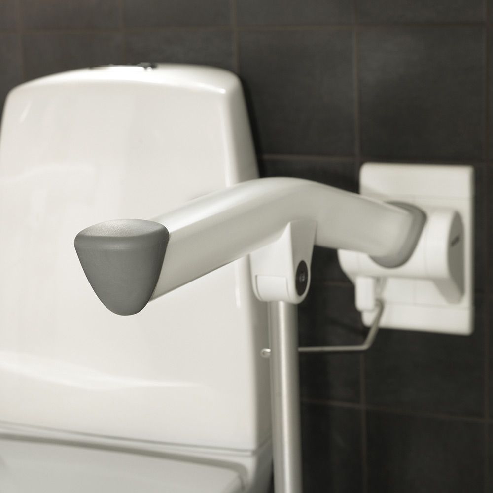Weißer Stützklappgriff mit grauem Griffende, neben einer Toilette montiert.