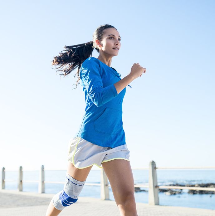 Frau beim Joggen mit Kniebandage. Die Bandage ist blau und grau. Sie ist am Knie getragen.