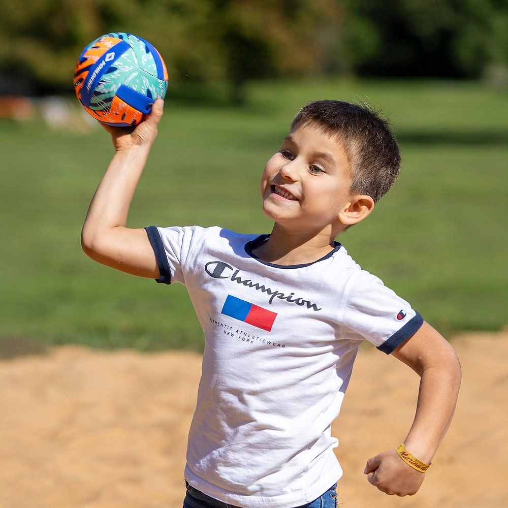 Kind wirft einen bunten Ball. Der Ball hat das Schildkröt-Logo. Kind trägt ein weißes T-Shirt.