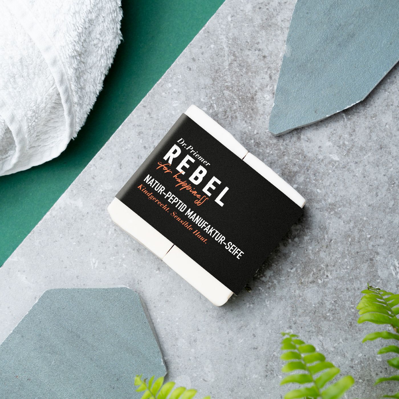 Seife mit schwarzem Etikett. Aufschrift: Dr. Priemer, REBEL for happiness, Natur-Peptid Manufaktur-Seife, kindgerecht, sensible Haut. Auf Stein.