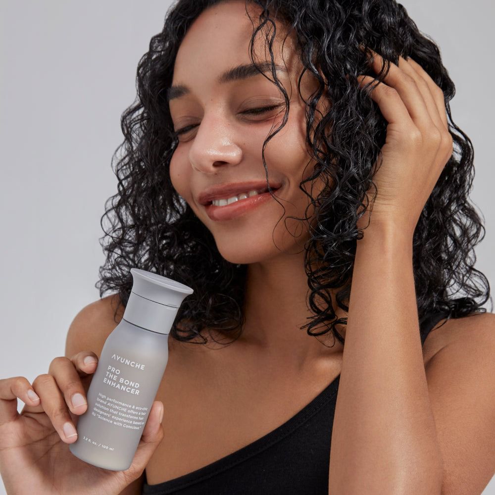 Frau mit lockigem Haar hält eine graue Flasche AYUNCHE PRO THE BOND ENHANCER.