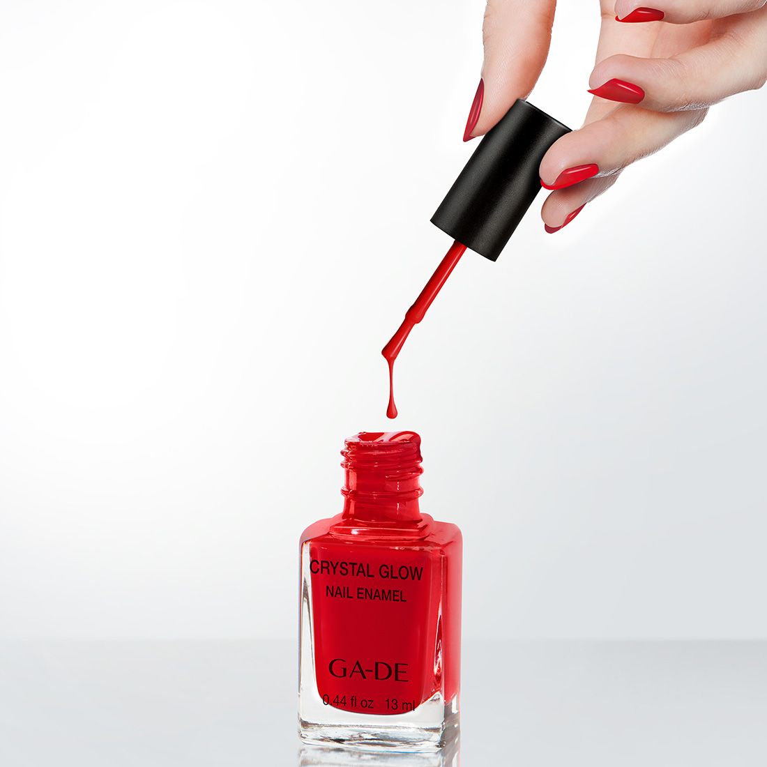 Roter Nagellack in Glasflasche, Pinsel mit Tropfen. Aufschrift: Crystal Glow Nail Enamel, GA-DE. Hand mit lackierten Nägeln.
