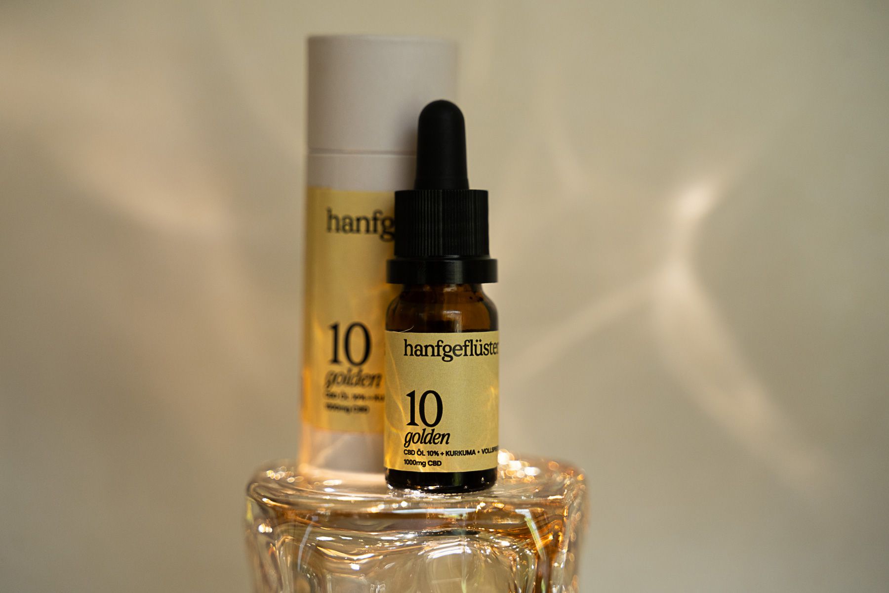 Braune Glasflasche mit Tropfer und zylindrischer Verpackung. Aufschrift: HANFGEFLÜSTER 10 golden.