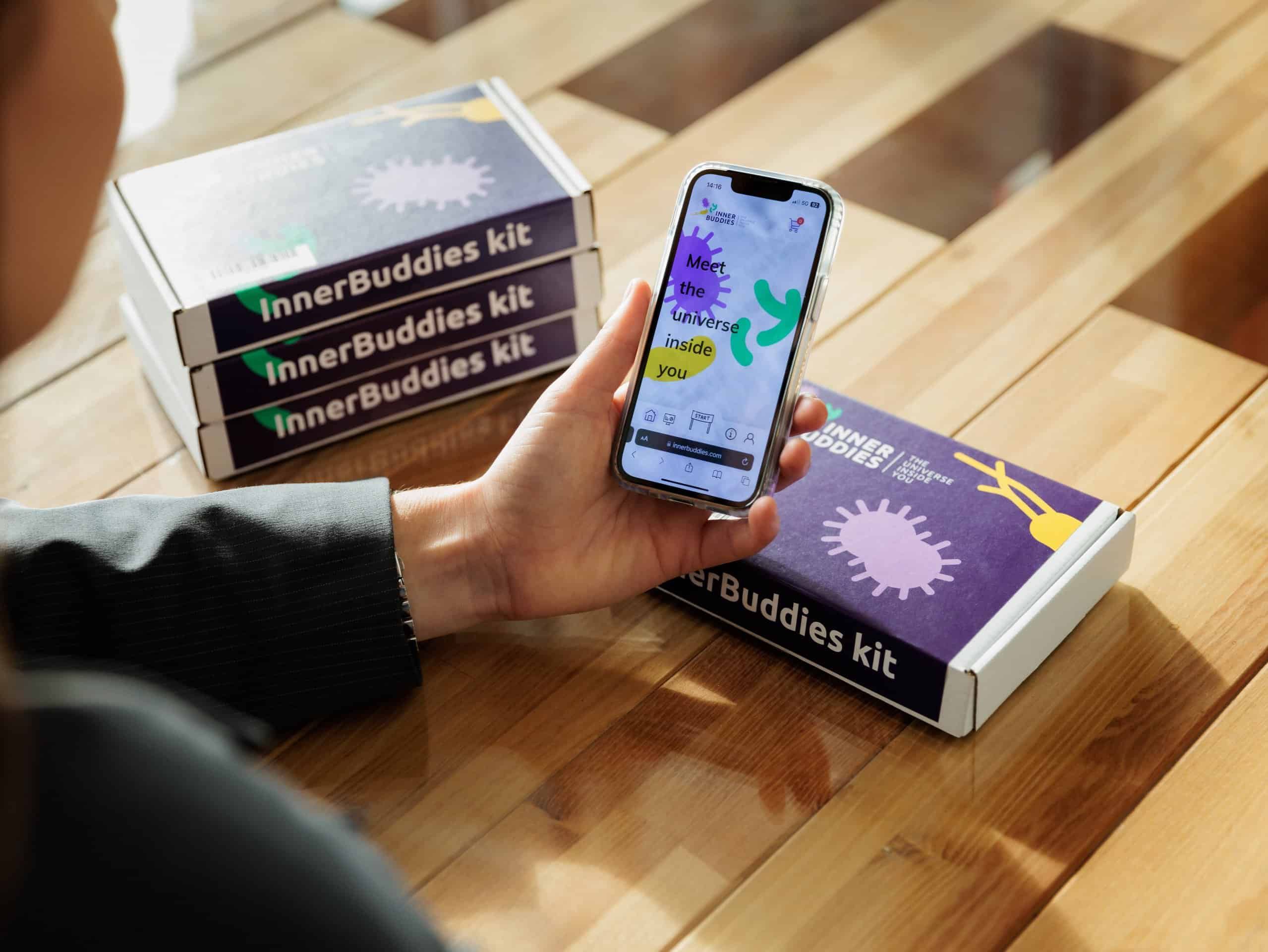 Drei gestapelte Inner Buddies Testkit-Verpackungen neben einer Person, die ein Smartphone hält.