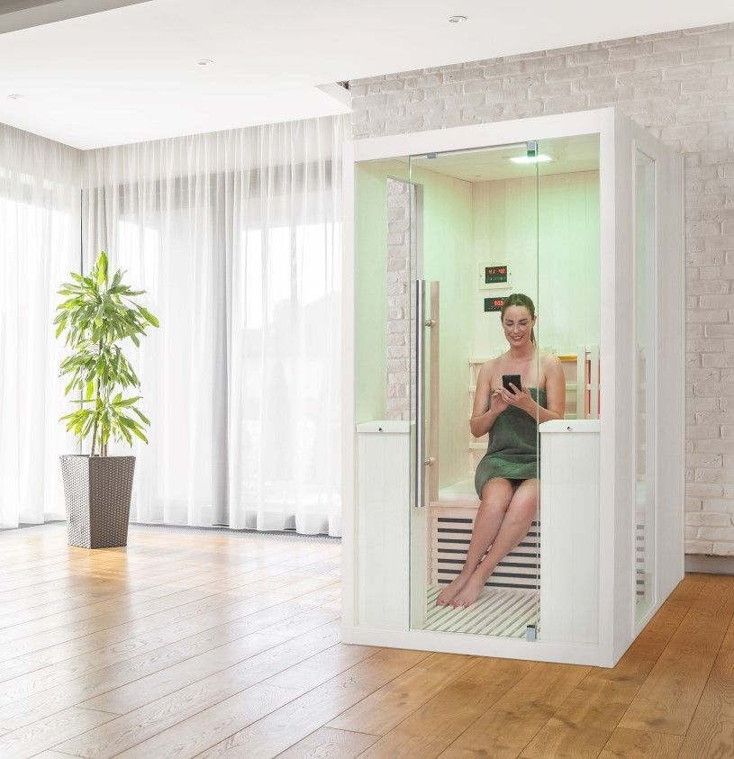 Infrarotsauna in hellem Raum. Frau sitzt in der Sauna, hält Smartphone. Sauna mit Glasfront und Holzelementen.