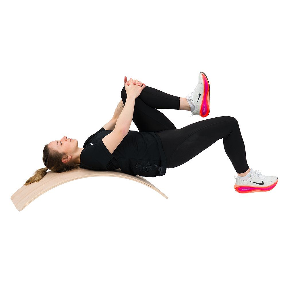 Frau liegt auf dem Rücken auf einem Balance Board, ein Knie zur Brust gezogen.