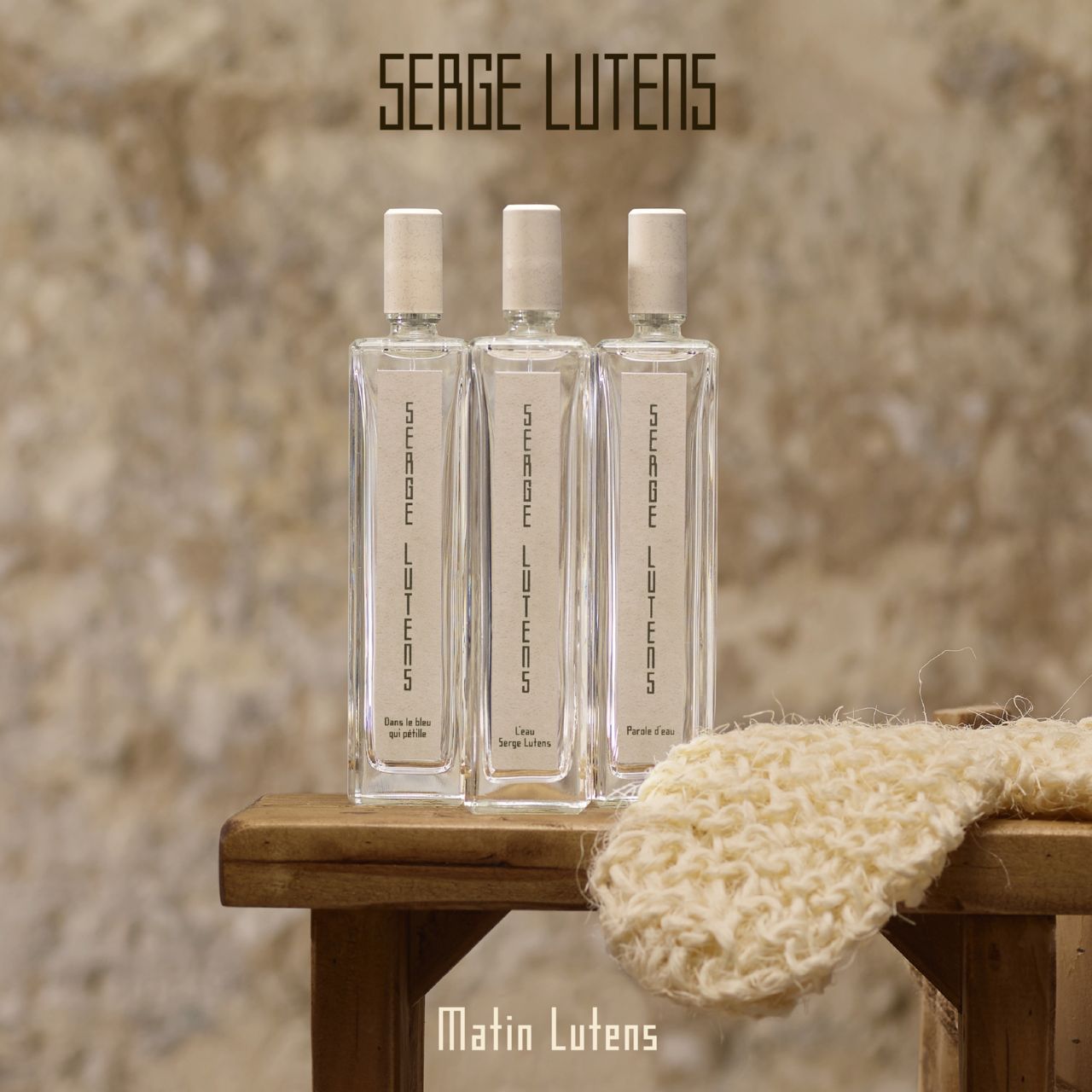 Drei rechteckige Glasflaschen auf einem Hocker. Aufschrift SERGE LUTENS. Unterhalb: Matin Lutens.