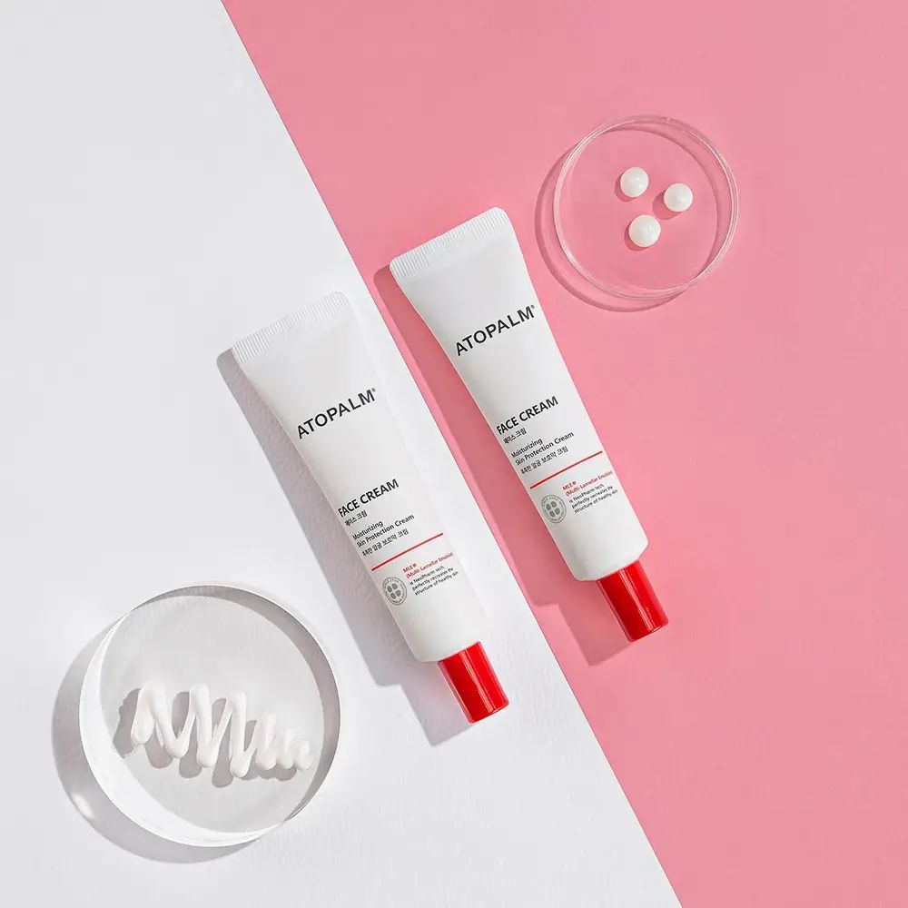 Zwei weiße Tuben mit rotem Verschluss. Aufschrift ATOPALM und FACE CREAM. Auf rosa und weißem Hintergrund.