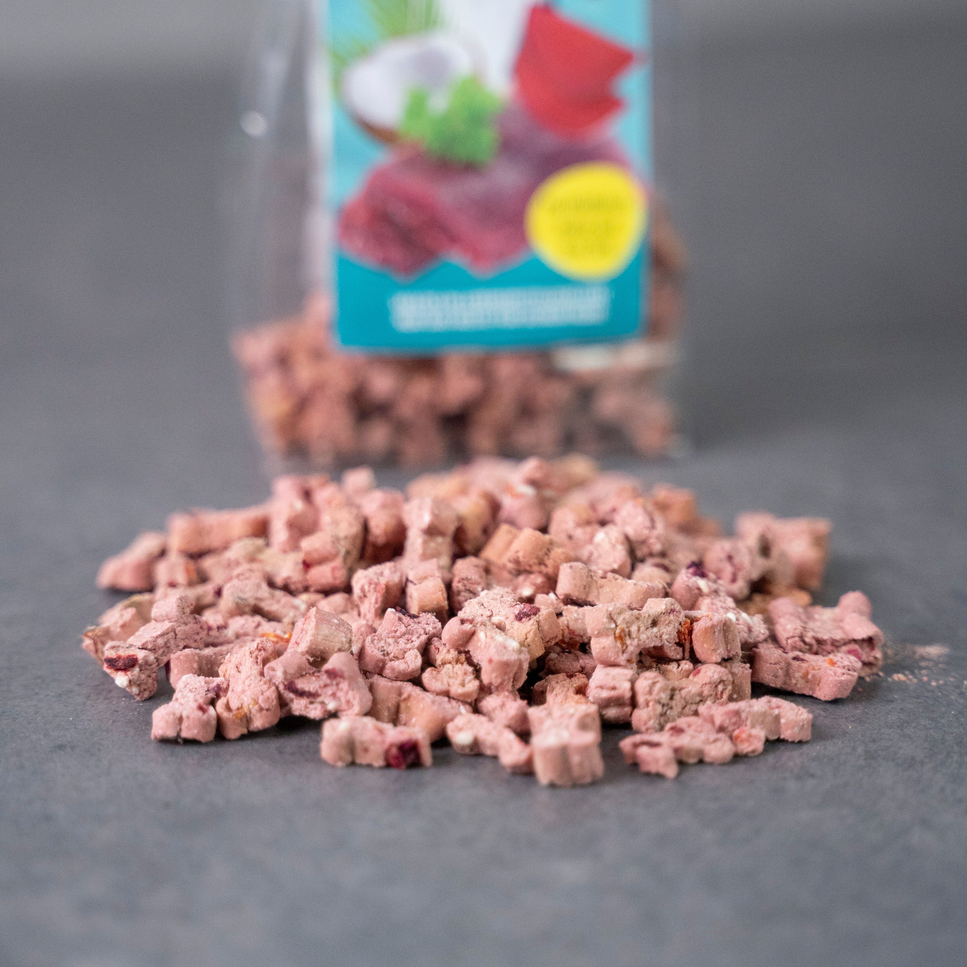 Haufen von rosa-braunen, würfelförmigen Hundesnacks. Im Hintergrund, teilweise unscharf, eine Tüte mit Etikett.