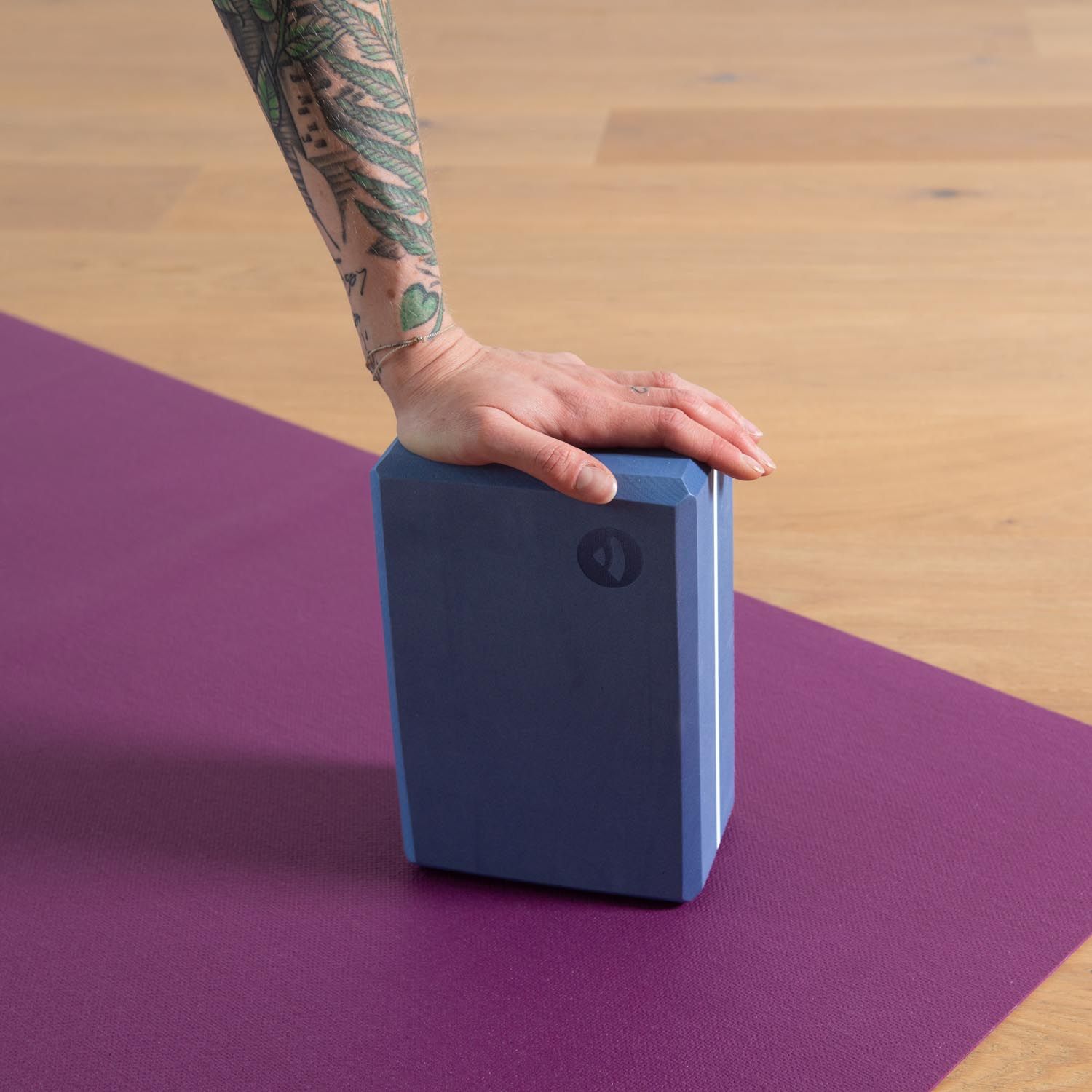 Hand auf blauem Yogabrick. Lila Yogamatte. Tattoo auf dem Arm. Helle Holzboden.