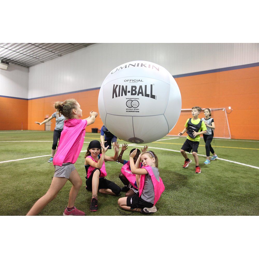 Großer weißer Kin-Ball. Kinder spielen in einer Sporthalle. Aufschrift: OMNIKIN, OFFICIAL KIN-BALL. Kinder tragen Westen.