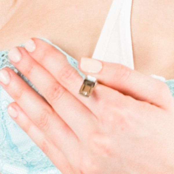 Nahaufnahme einer Hand, die einen Ring mit einem Stein trägt. Im Hintergrund ist ein hellblaues BH-Oberteil zu sehen.