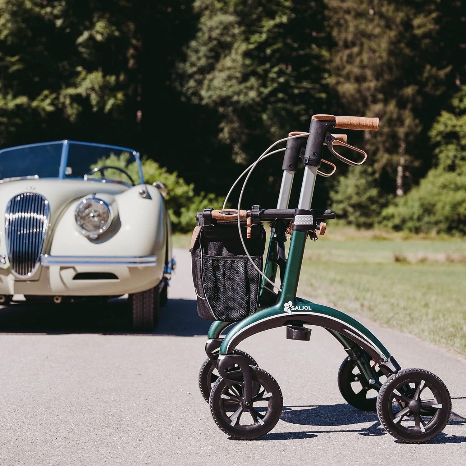 Grüner Rollator auf Asphalt, im Hintergrund ein Oldtimer-Cabriolet. Griffe und Tasche sind sichtbar. Bäume im Hintergrund.
