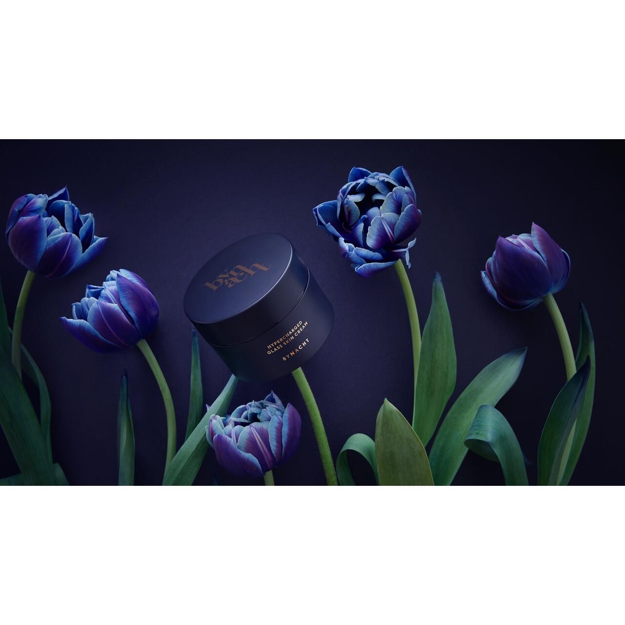 Dunkelblauer Tiegel, umgeben von blauen Tulpen. Aufschrift: Hypercharged Glass Skin Cream, Bynacht.