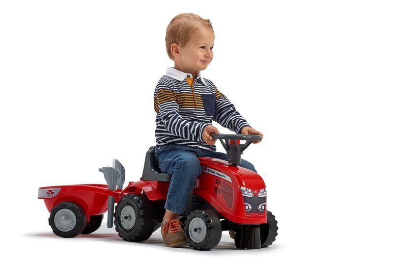 Kind sitzt auf rotem Spielzeugtraktor mit Anhänger. Der Traktor hat schwarze Räder und das Logo Massey Ferguson. Ein Kind sitzt auf dem Traktor.