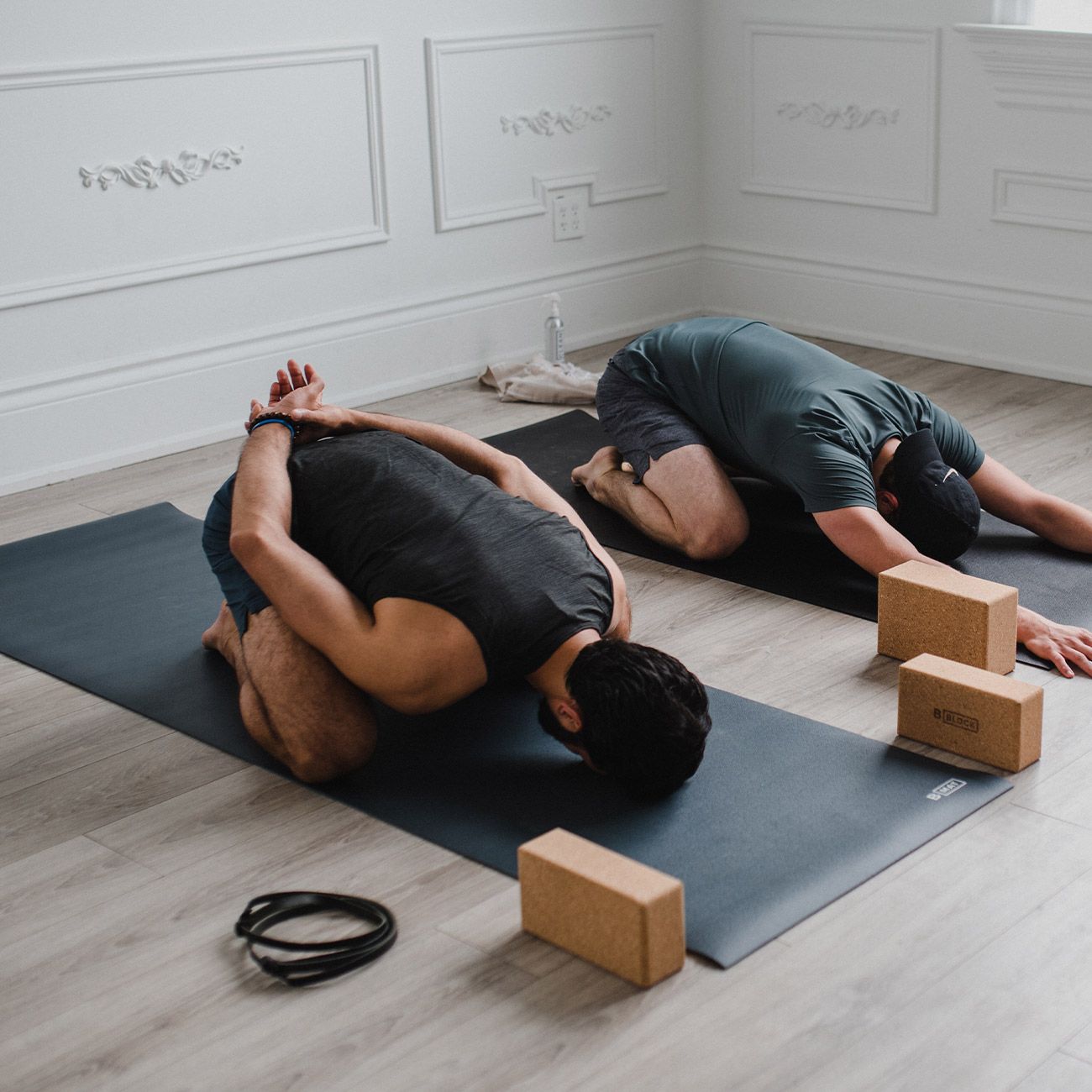 Zwei Personen praktizieren Yoga auf anthrazitfarbenen Matten. Yogablöcke und ein Gurt liegen daneben.