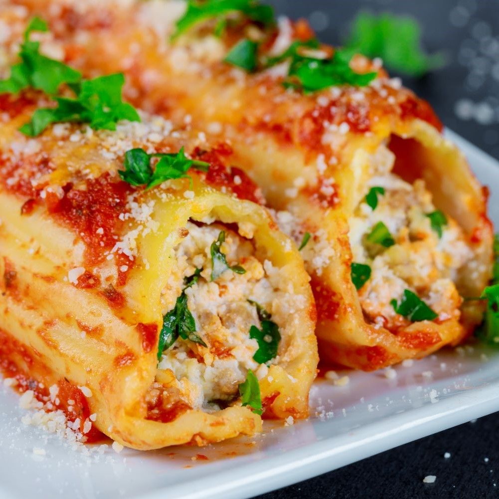 Nahaufnahme von drei Cannelloni-Rollen mit Füllung, bedeckt mit Tomatensauce und Kräutern. Auf weißem Teller.