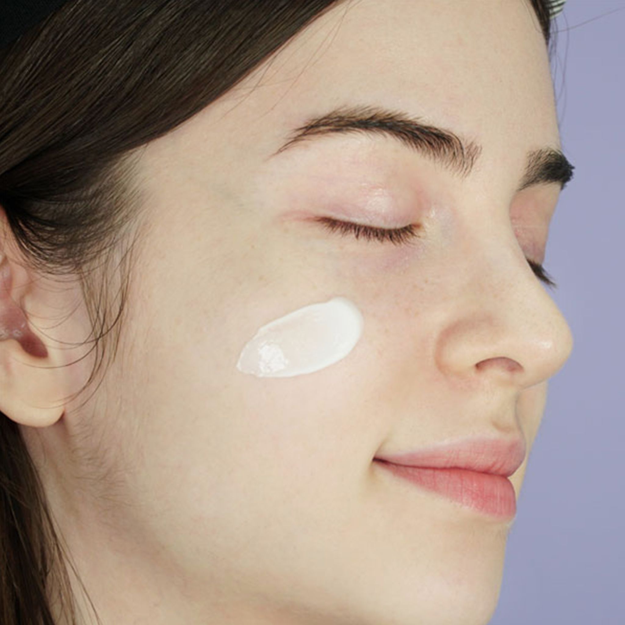 Gesicht einer Frau mit geschlossenen Augen. Auf der Wange ist eine Creme aufgetragen.